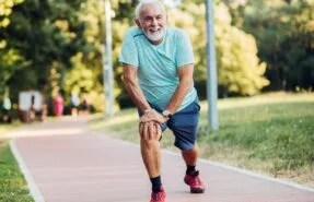 Una persona mayor, con una sonrisa en el rostro, trotando o caminando en un parque soleado, transmitiendo energía y bienestar. Podría estar acompañada de otra persona o sola, mostrando que la actividad física es posible y agradable en la tercera edad.