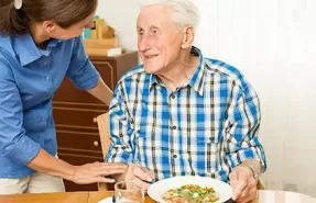 Adulto mayor disfrutando una comida saludable en casa, acompañado por un nutricionista.