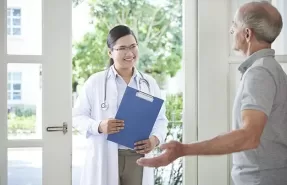 Una persona mayor sonriendo y relajada en el sofá de su casa mientras un profesional de la salud la atiende amablemente, ilustrando la comodidad y el apoyo emocional de la atención médica a domicilio.