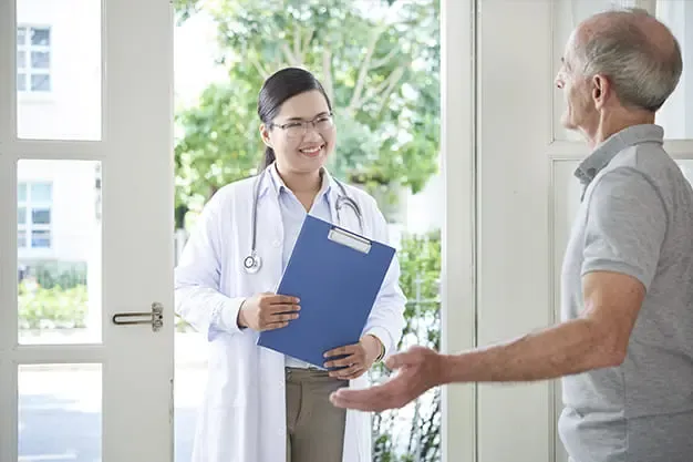 Una persona mayor sonriendo y relajada en el sofá de su casa mientras un profesional de la salud la atiende amablemente, ilustrando la comodidad y el apoyo emocional de la atención médica a domicilio.