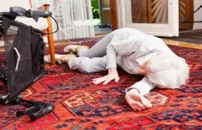 Una adulta mayor sentada en el suelo de su casa con una expresión de dolor o sorpresa tras una caída, con un objeto que pudo causar el tropiezo cercano.